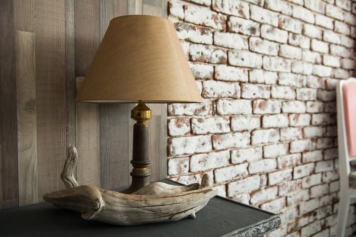 A wooden lamp with a beige shade sits on a dark table beside a brick wall.