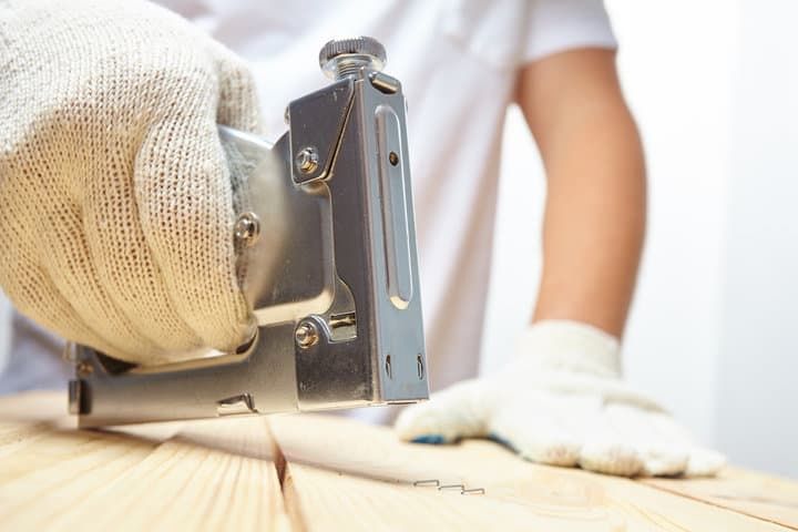 A gloved hand operating a staple gun on wooden planks.