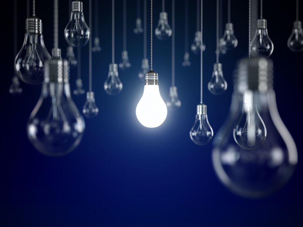 A glowing bulb shines among many hanging light bulbs against a dark background.