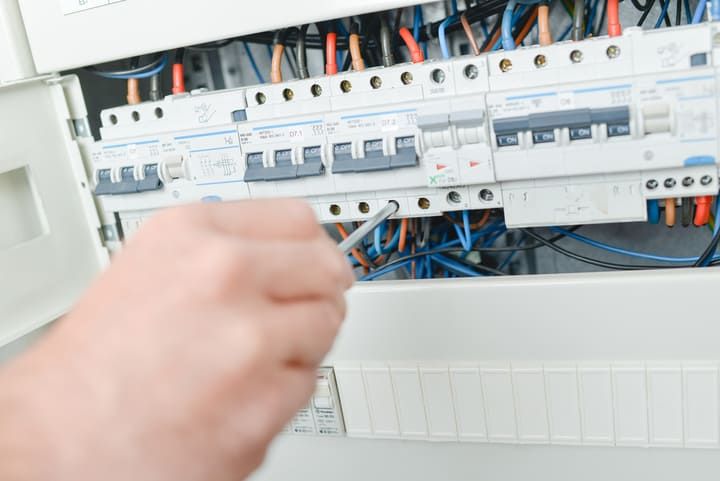 A hand uses a screwdriver on a circuit breaker panel with colored wires.