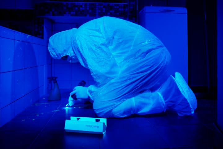 Person in protective suit kneeling, inspecting a surface under blue lighting.