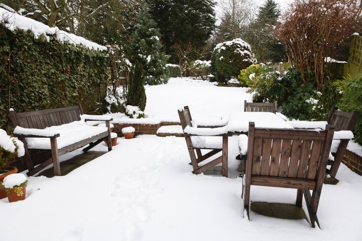 Snow-covered garden with wooden benches and tables, surrounded by green hedges.