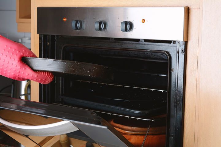 A hand wearing a heat-resistant glove removes a baking tray from an oven.
