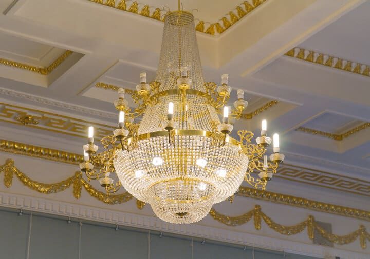 A grand chandelier with crystal elements and multiple light bulbs, hanging from a decorated ceiling.