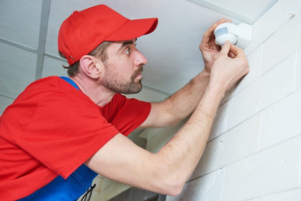 A technician installs a motion sensor on a ceiling near white tiled walls.