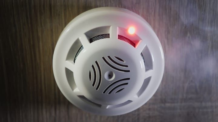 A white smoke detector mounted on a wall, featuring a glowing red indicator light.