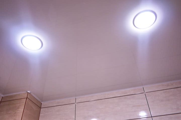 Two round LED ceiling lights illuminating a smooth ceiling and wall tiles.