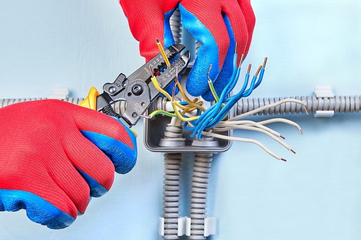 Hands in gloves using wire cutters on colored electrical wires in a junction box.