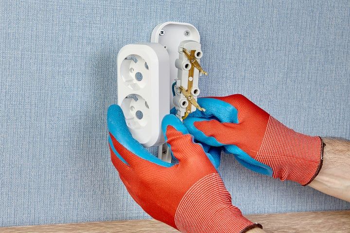 Hands wearing red gloves installing an electrical outlet on a blue wall.