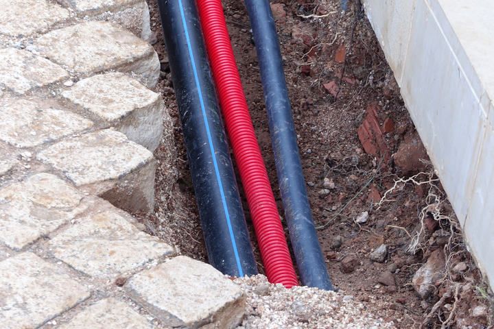 Trenches showing black and red pipes alongside paved stones and soil.