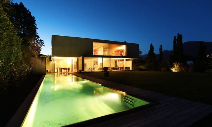 Modern house with illuminated pool reflecting evening sky and surrounding trees.