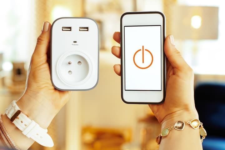 A person holds a smart plug and smartphone displaying a power button icon.