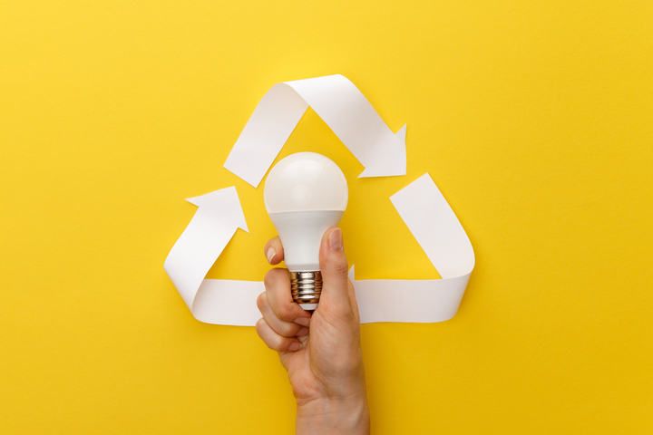 A hand holding an LED light bulb surrounded by recycling arrows on a yellow background.