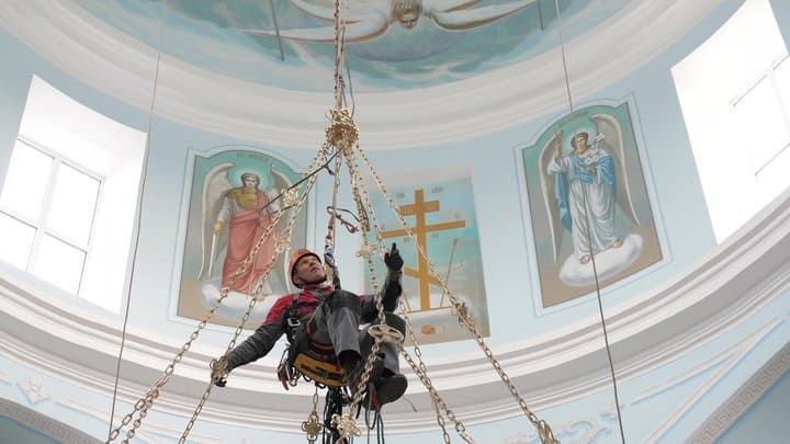 A worker suspended from a ceiling, repairing lighting in a church with angelic murals.