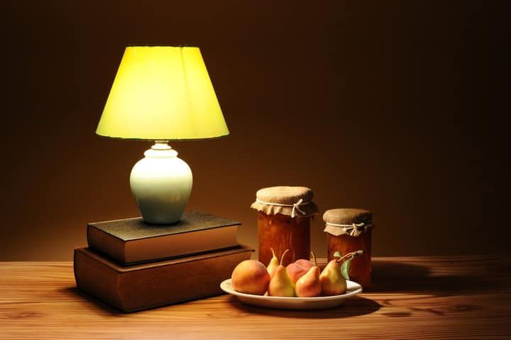 A lit lamp beside books, jars of jam, and fruit on a table.