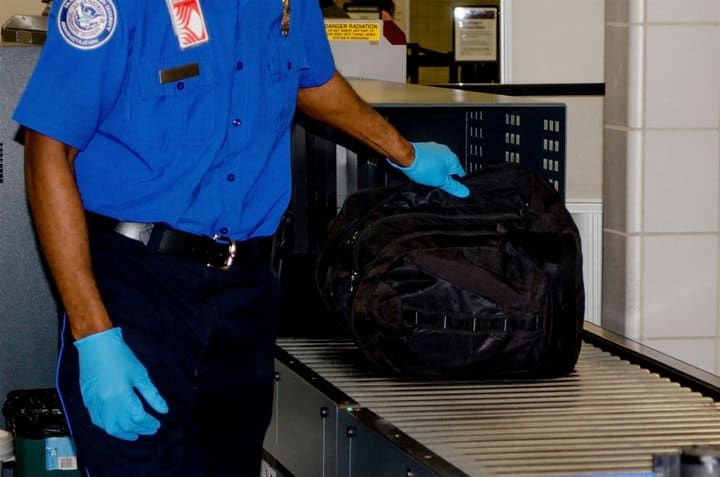 TSA agent wearing gloves inspects a black bag on a conveyor belt.
