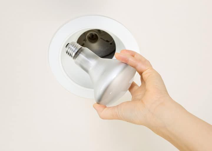 A person holding a silver LED bulb near a ceiling fixture.