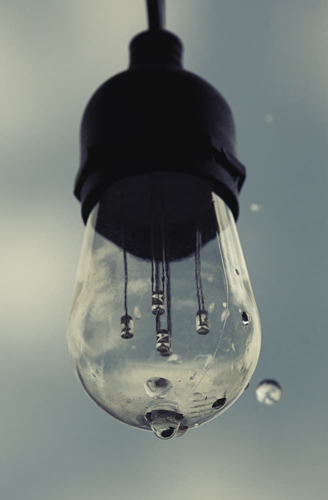 A hanging LED bulb with droplets on its glass, illuminating a cloudy background.