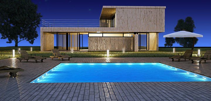 Modern wooden house at night with illuminated pool and lounge chairs.