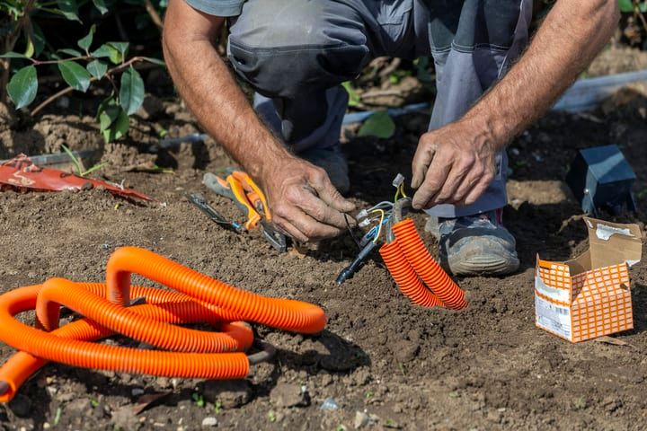 A person connects electrical wires near coiled orange tubing on soil.