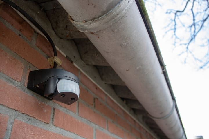 A motion sensor light mounted on a brick wall under a metal gutter.