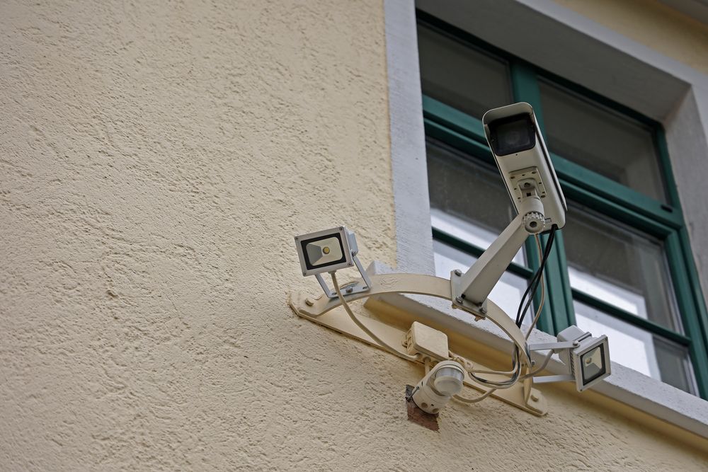 A wall-mounted security camera with LED floodlights installed beside a window.