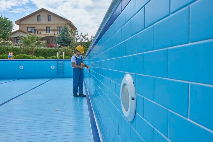 A worker in a yellow hat inspects a bright blue tiled swimming pool wall.