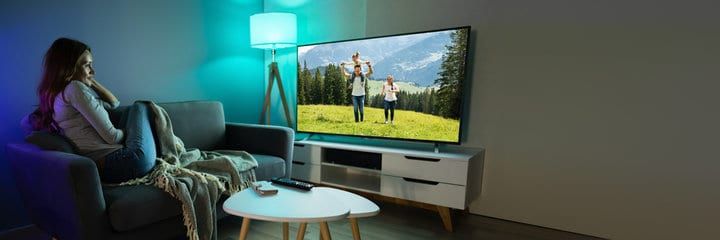 A woman relaxes on a couch watching a bright television scene of a family outdoors.