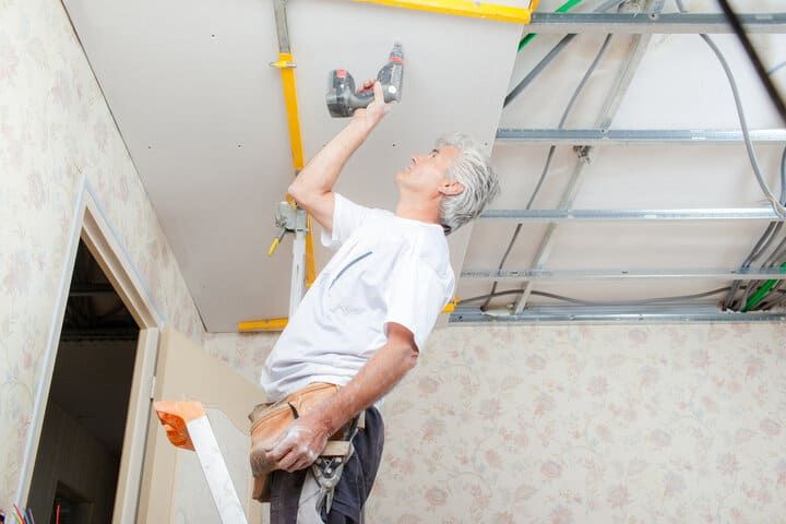 A man uses a power drill while standing on a ladder, installing ceiling lighting.