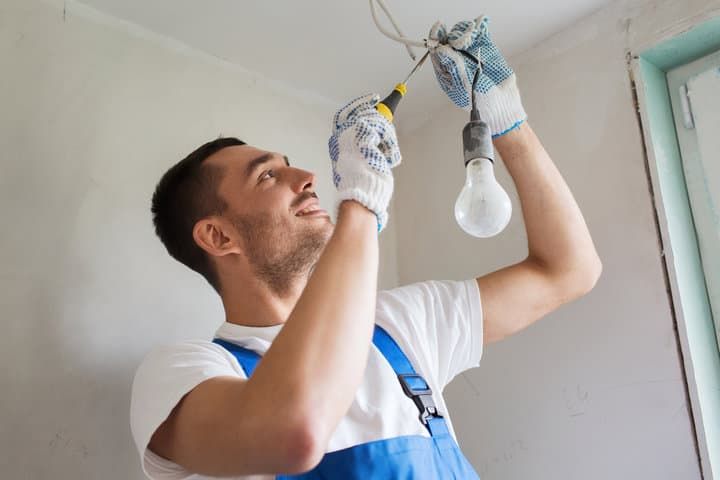 A worker installs a light bulb in a ceiling fixture with tools.