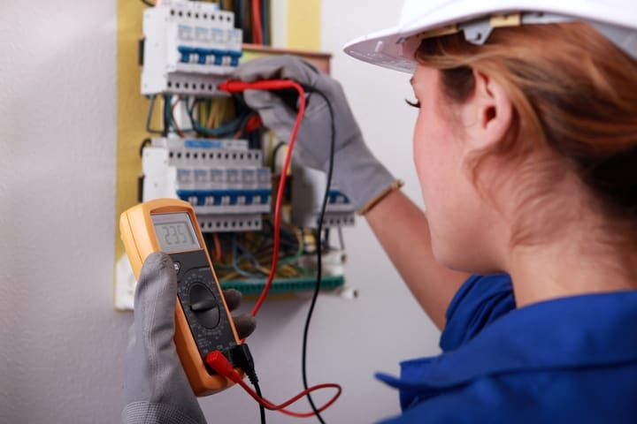 An electrician uses a multimeter on electrical panels while wearing gloves and a hard hat.