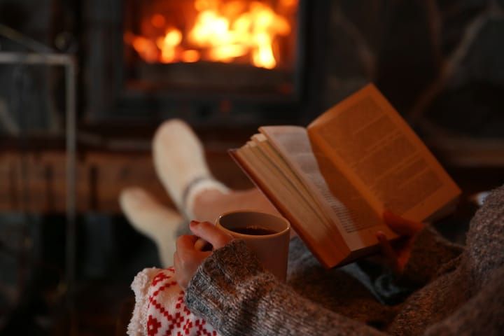A person reads a book by a cozy fireplace, holding a warm beverage.