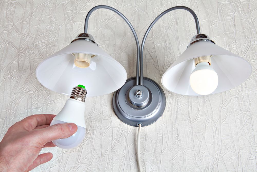 A person holds a new LED light bulb near a wall-mounted lamp.