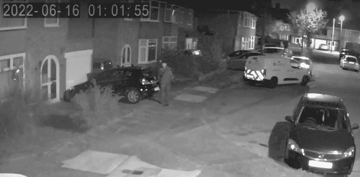 A nighttime scene showing a man near a parked car on a quiet street.