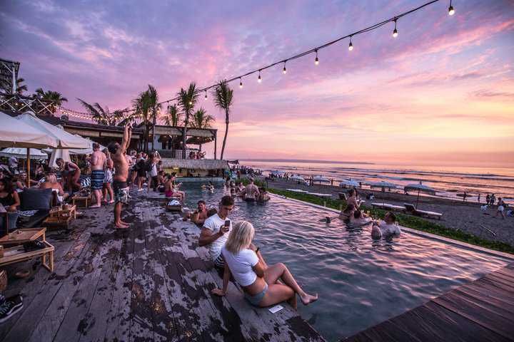 People enjoying a vibrant sunset at a beachside pool with decorative lights.