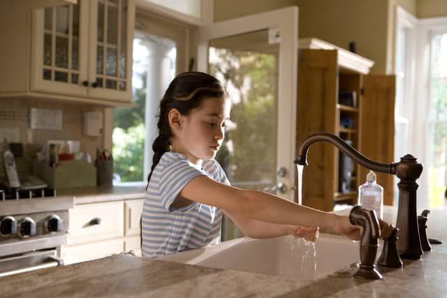 A young girl washing her hands at a kitchen sink under natural light.