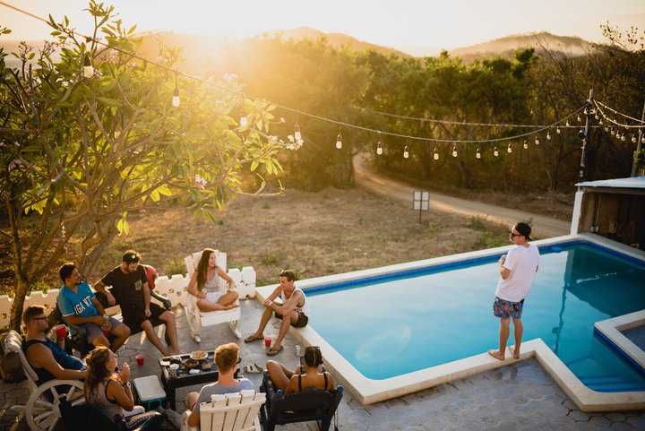 Group of friends gathered around a pool during sunset, with string lights above.
