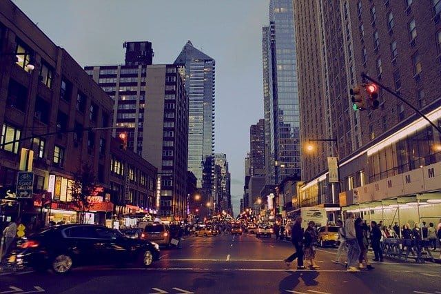 Busy city street at dusk with cars and pedestrians among tall buildings.